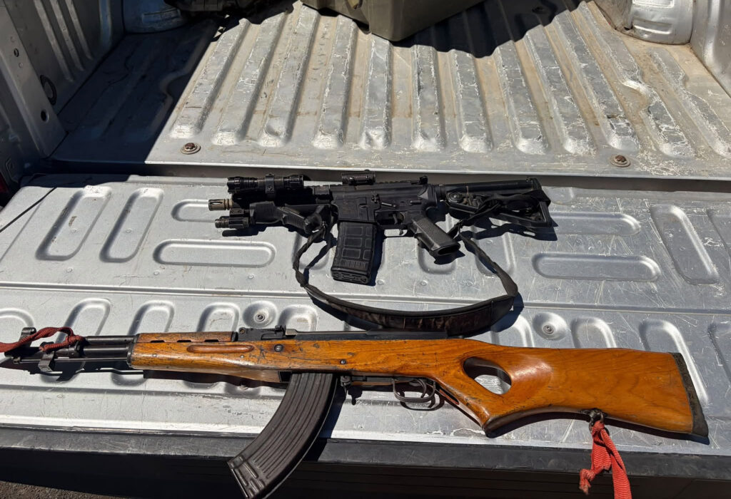 Two rifles, one modern black assault rifle with accessories and one older wooden-stocked rifle, rest on the metallic bed of a pickup truck.