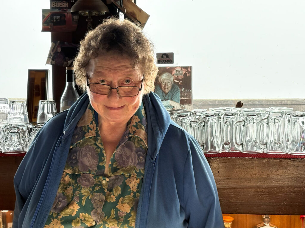 An older person with curly hair and glasses smiles while standing behind a bar counter filled with glassware.
