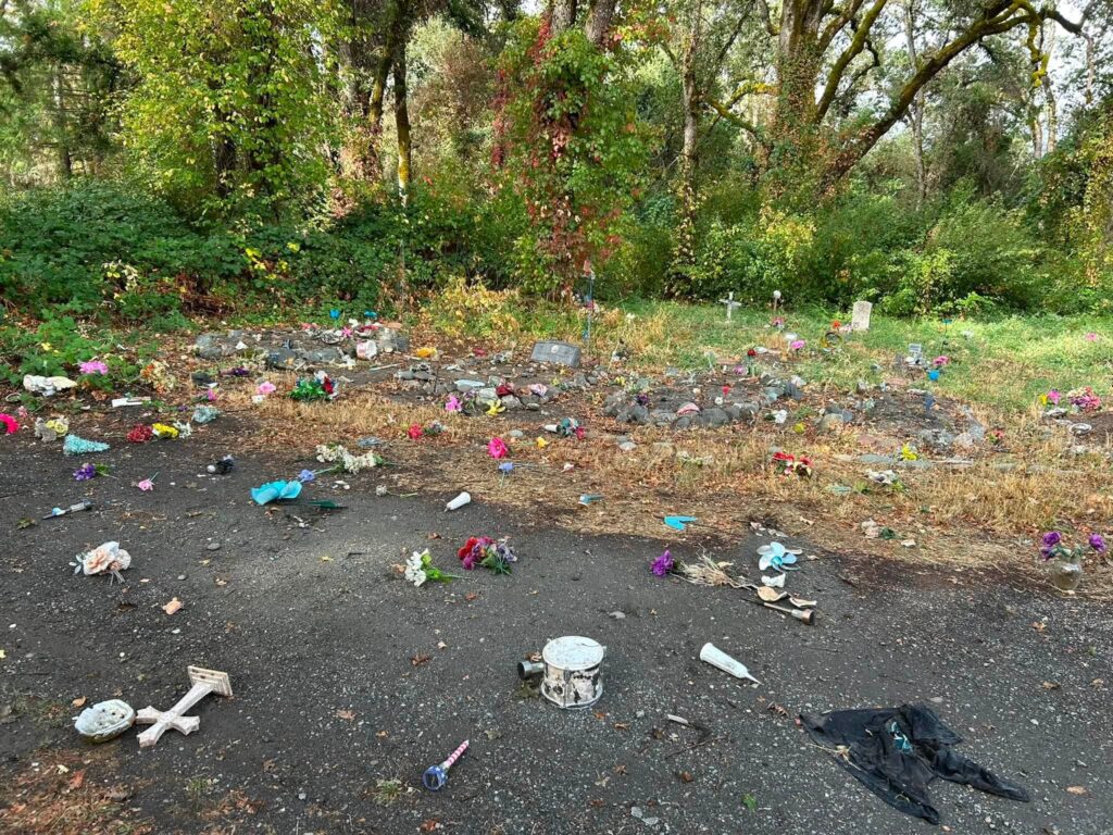A makeshift memorial site outdoors, scattered with flowers, crosses, and various items on dirt and gravel ground, surrounded by trees.